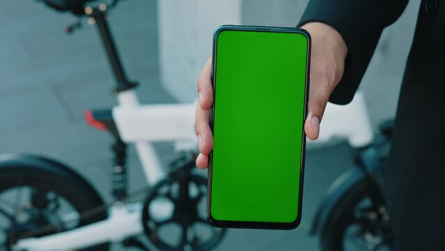 Male hands holding smartphone with green screen on the electric bike background. Social media, online shopping mockup with mobile phone device. Man showing the green screen.