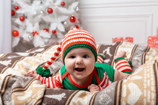 Newborn baby girl dressed in gnome costume lying on white fur carpet among christmas decorations. Christmas photo of infant in ctriped cap. New Year concept.