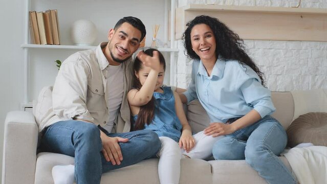 Multiracial family multiethnic Hispanic Caucasian Arabian Indian parents mother father with daughter child girl sitting on couch at home waving hands hello talking on online video call chat conference