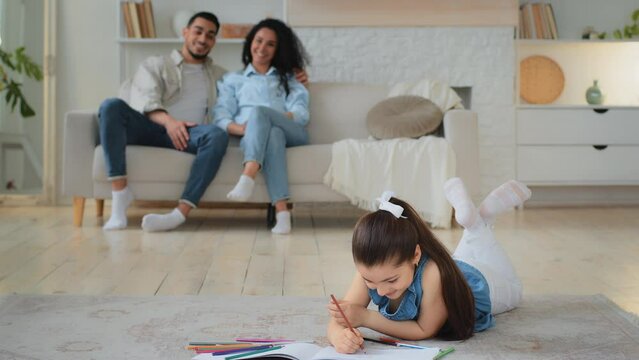 Little Caucasian daughter schoolgirl girl kid lies on floor draws picture on paper with colored pencils drawing painting hobby multiracial multiethnic parents mother and father couple sitting on sofa