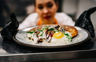 Chef's hands in gloves cooking fried pancake with mushroom sauce, bacon and egg on kitchen