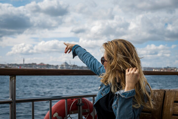 Blonde long-haired woman traveling by ferry in Bosphorus, pointing to a point with her hand.