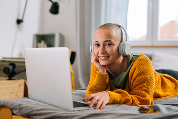 teenager shaved head female with headphones laying on bed in her room and using laptop