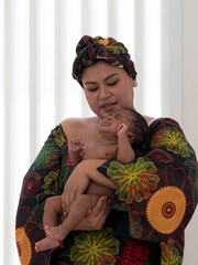 Asian mother wearing traditional African clothes with headscarf holding and calming crying cute little mixed racial newborn baby boy.