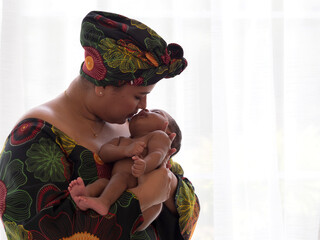 Asian mother wearing traditional African clothes with headscarf kissing cute little mixed racial newborn baby boy.
