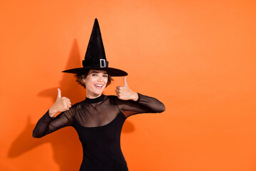 Photo portrait of gorgeous young lady show double thumb up smile wear stylish black halloween witch clothes isolated on orange background