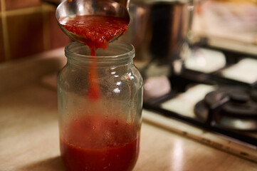 Cropped view of a housewife in black chef's apron using a ladle, pours boiling tomato juice from saucepan into sterilized jar while cooking homemade passata in the kitchen at home. Canning food