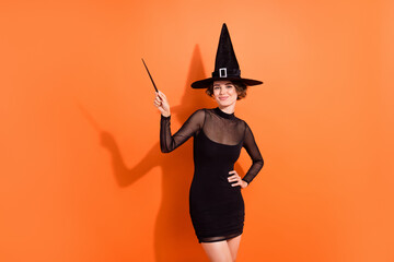 Photo of cute young woman point wooden wand hogwarts theme event dressed trendy black halloween witch look isolated on orange background - obrazy, fototapety, plakaty