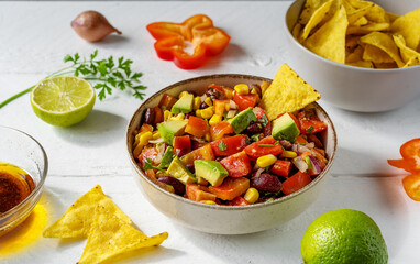 Cowboy caviar is traditional Mexican vegetable salad in bowl with ingredients and nachos on white wooden table with ingredients