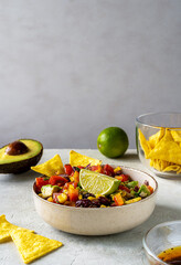 Cowboy caviar is traditional Mexican vegetable salad in bowl with ingredients and nachos on grey background