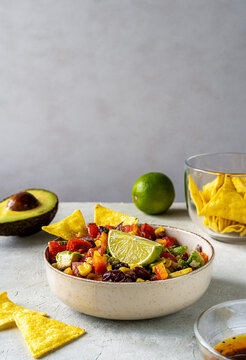 Cowboy caviar is traditional Mexican vegetable salad in bowl with ingredients and nachos on grey background