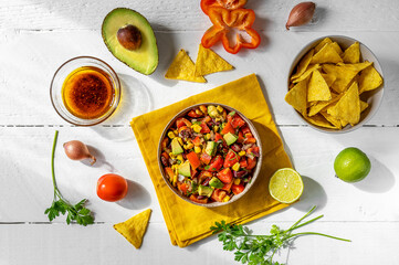 Traditional Mexican vegetable salad . cowboy caviar in bowl with ingredients and nachos on white wooden table with ingredients