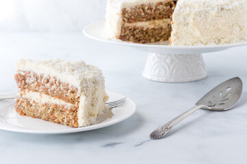 A white frosted coconut covered carrot cake with one slice ready for eating.