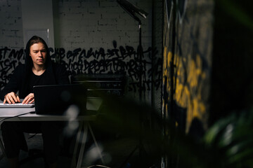 Focused hacker man in sweatshirt hacking online web site or engaging hacking into security systems, using laptop and desktop computer at desk in dark room. Concept of data thief, internet attack.