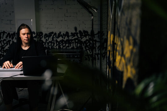 Focused hacker man in sweatshirt hacking online web site or engaging hacking into security systems, using laptop and desktop computer at desk in dark room. Concept of data thief, internet attack.