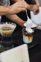 Barista making iced latte coffee with hand topped whipping in cafe.