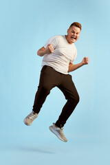 Fun, joy. Tall teen boy, overweight model in white shirt shouting and jumping isolated over blue...