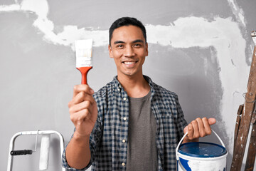 An attractive Asian man holds up paintbrush, tin and smiles before painting