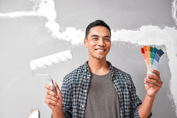 A young Asian man holds paint swatches and roller about to paint repaired wall