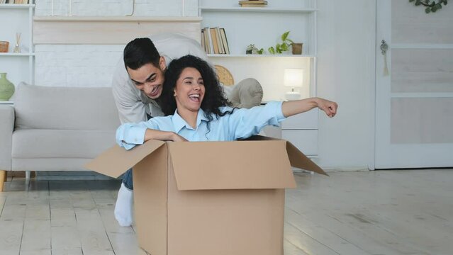 Hispanic Indian man husband boyfriend pushing large cardboard box with wife funny woman sitting inside riding in parcel married couple celebrating relocation buying new house real estate property loan