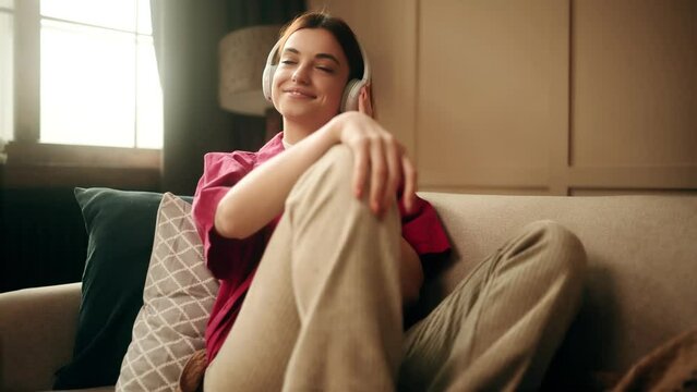 Charming young woman relaxing on comfortable sofa wearing headphones. Pretty lady enjoys listening chill music at home.