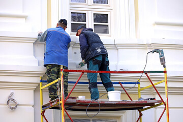 Workers repair the building wall standing on lifting platform. Two builders painting the house...