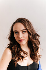 Close-up portrait of brunette woman. Curly blue-eyed girl in black tee smiles and looks into camera on white background