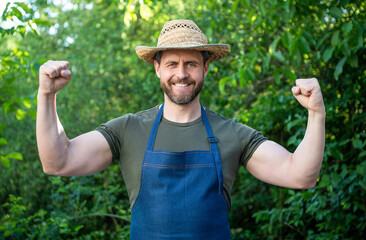 Happy strong farmer in farmers hat and apron flexing arms on farm outdoors