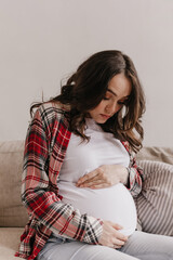 Brunette attractive pregnant woman looks and gently touches belly. Portrait of happy young mother sitting on sofa