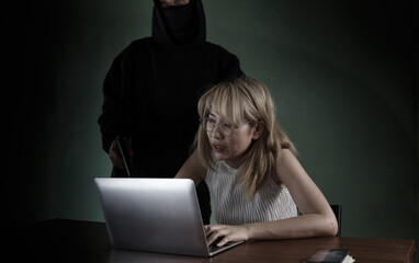 Fototapeta premium A black robed criminal with a gun at her aimed at a woman to steal her computer information. in a pitch dark room
