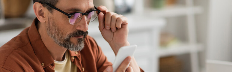 man with grey beard looking at smartphone while touching eyeglasses, banner.