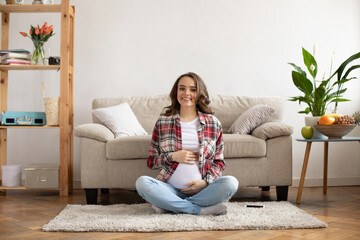 Cheerful brunette pregnant woman in red plaid shirt sincerely smiles and gently touches belly. Happy young mother sitting on soft beige carpet in light living room