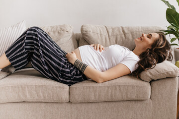 Happy curly woman in striped black pants and white tee lies on beige sofa and smiles. Pregnant lady gently touches belly