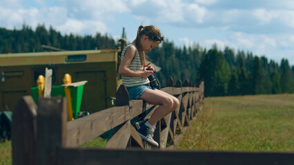 Young photographer wiping digital camera lens by t-shirt, sitting on wooden fence. Girl shooting beautiful nature landscape, concentrating on process, spending leisure time outdoor. Slow motion.