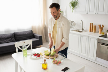 smiling man cutting lettuce near bowl with vegetable salad and bottles with oil.