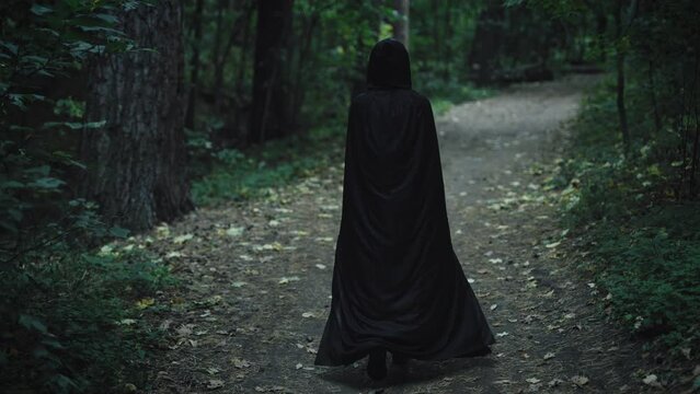 A dark figure in a black cloak with a hood walks through the enchanted forest. An unrecognizable witch from the Middle Ages walks along the path.