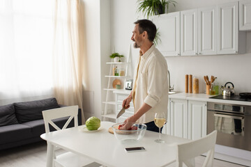 happy bearded man looking away while standing near fresh vegetables and smartphone with blank screen in kitchen.