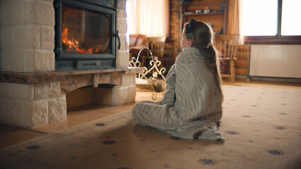 Young girl sitting by the fireplace on mild carpet, watching on fire and warming by coverlet in...