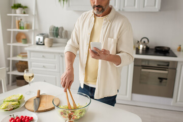 partial view of man preparing salad with fresh lettuce and cherry tomatoes while looking at cellphone.