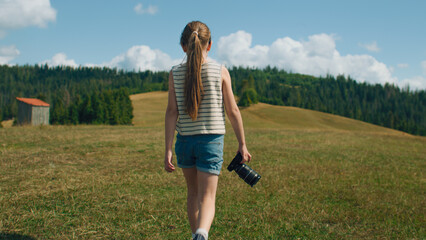 Young photographer walking with camera on grass hill. Girl watching at beautiful landscape and amazing nature view, spending leisure time outdoor. Slow motion. Back view.