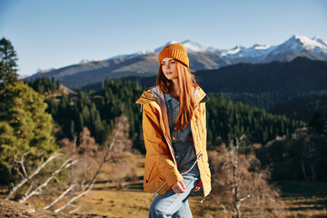 Naklejka premium Woman hiking in the mountains in the fall with a smile with teeth and happiness in a yellow cape with red hair full-length stands against the backdrop of trees and mountains in the sunset light