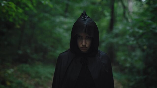 Portrait of a witch in a black cloak with a hood. Woman in the mystical forest. Dark forces