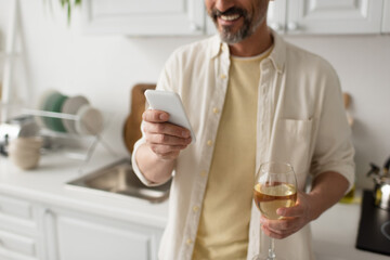 partial view of smiling bearded man with glass of white wine using smartphone in blurred kitchen.
