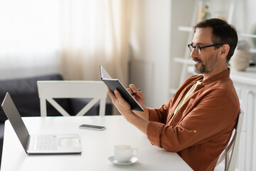 side view of smiling man in eyeglasses writing in notebook near laptop and coffee cup.