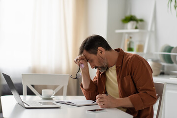 exhausted man sitting with bowed head near devices and empty notebook in kitchen.