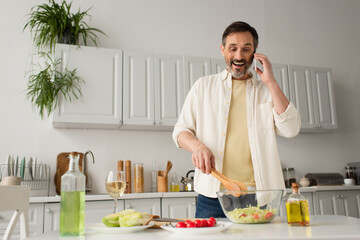 Fototapeta na wymiar cheerful man preparing vegetable salad while talking on cellphone in kitchen.