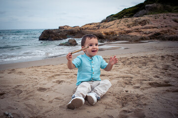 Niño pequeño bebé jugando con un palo en la mano, sobre la arena de la playa con el mar de fondo 