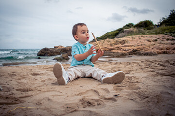 Niño pequeño bebé jugando con un palo en la mano, sobre la arena de la playa con el mar de fondo 