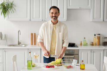 bearded man smiling at camera while preparing salad with lettuce and cherry tomatoes near glass of white wine.