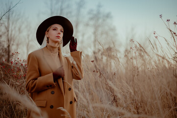 Elegant fashionable blonde woman wearing trendy autumn outfit with classic brown coat, hat, suede gloves, beige turtleneck, golden earrings, posing outdoor. Copy, empty space for text